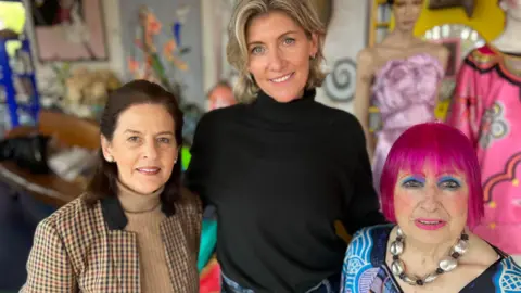 A woman on the left is wearing a brown checked blazer, a woman is wearing a black turtle neck jumper and a woman wearing a blue multi-coloured shirt. They are standing together.