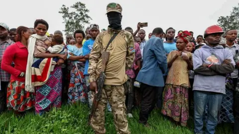 EPA stands in front of a crowd of citizens with a member of M23 in a Balaclava and a automatic weapon.