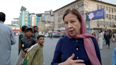Two boys; out of focus, stare at Lyse Doucet wearing a pink headscarf.