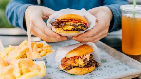 A man holding a cheeseburger in paper packaging, only his hands are visible. Another burger sits on a metal tray, with two portions of chips in cardboard trays, and an orange soft drink in a clear cup seen to the side. 