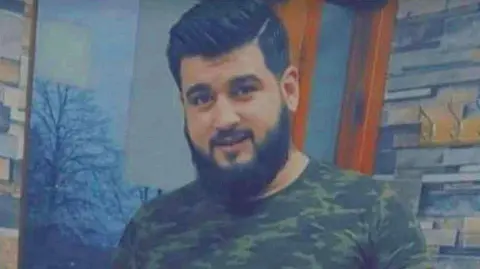 Anas Almarjeh smiles into the camera - he is wearing a T-shirt with military camouflage, while standing indoors. He has dark hair and a beard.