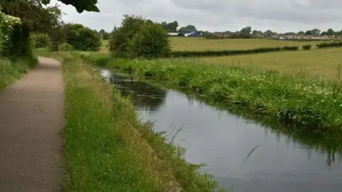 NChadwick/Geograph A path runs alongside a river that has formed in the remnants of the Manchester, Bolton & Bury canal. A bird can be seen wading in the river, which is flanked on the opposite side with green fields, with houses seen in the far distance.