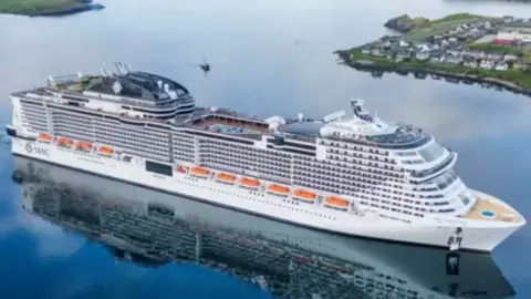 David Donaldson/Shetland Flyer Aerial Media A white cruise ship with multiple decks and flanked by orange lifeboats on a calm inlet with a pool visible on the top deck with a background of a spit of land on either side. 