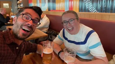 John Robinson/Claire Reynolds The couple from the above pictures sitting in what looks like a restaurant, both smiling at the camera. They are drinking pints of beer. The man is wearing a red check shirt and the woman is wearing a white t-shirt with a dark blue and a light blue stripe across it.