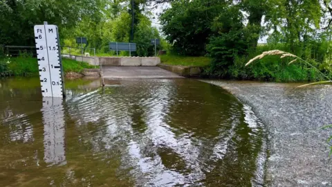 Martin Giles/BBC A view of Shotesham ford. It shows a large body of water moving over the road. Barricades can be seen at the end of the road while a water gauge sits on the left hand side showing the depth of the water.