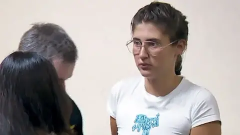 Rustavi 2 Bella Culley filmed in court. She is wearing glasses and has brown hair in a plait and is wearing a white top 
