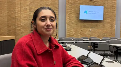 Martin Heath/BBC Shanika Mahendran with long dark hair tied back, wearing a red coat and sitting in a council chamber with white tables, and microphone units. There are grey seats behind the tables and brick walls in the background.