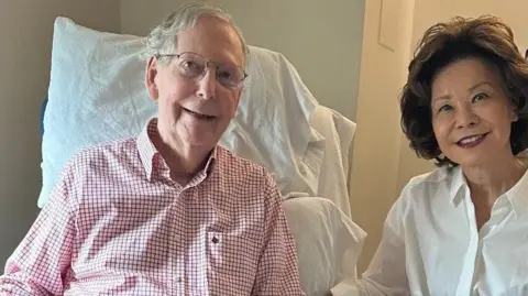 Mitch McConnell sits looking ahead in red checkered shirt, next to his wife who is wearing a white shirt