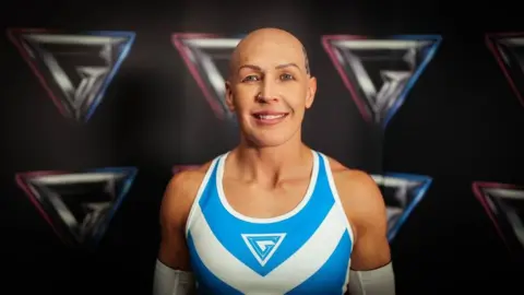 BBC/ Hungry Bear Media Ltd/ David MacCormack A white, female competitor wears the official 'blue team' kit and smiles happily in front of a Gladiator-branded backdrop.  She has a bald head, wears a little makeup and looks fit and muscular.