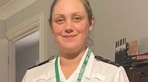 Ministry of Justice A smiling blonde woman in a white prison officer uniform and wearing a green lanyard.