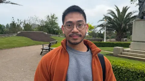 Emmanuel Delmoro is wearing an orange jacket, grey top and glasses. He has black hair and is pictured in Howard Davis Park