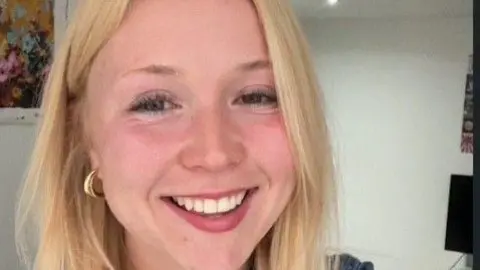 Aneesa Aneesa is a young woman with long blonde hair worn down. She smiles at the camera and wears a denim top and gold necklace and earrings