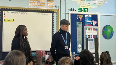 Two sixth-form students stand in front of a whiteboard and posters at the front of a classroom. A row of pupils are sat in front.