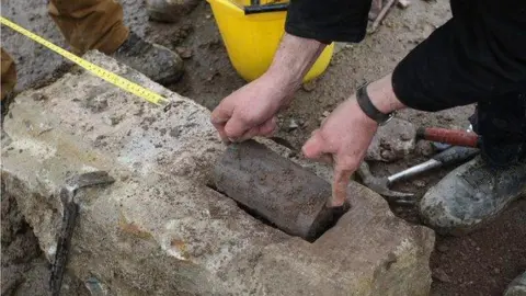 DCSDC a lead capsule is removed from rock by archaeologists. a pair of hands is seen lifting it the capsule out of the rock. a tape measure is seen in the background, as are a workers boots.