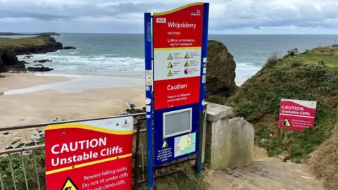 BBC Three signs stand on the top of a cliff with Whipsiderry beach behind and below them. The red warning signs detail the dangers of unstable cliffs and rock falls and have emergency contact details for use in case of an accident.