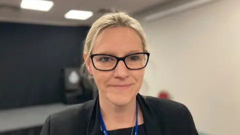 BBC/Laura Coffey Woman looking at camera wearing black glasses a black suit jacket and blue lanyard