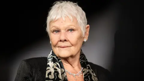 Getty Images Dame Judi Dench, a woman with short grey hair, wears a dark jacket and black and white silk scarf, as she looks directly into the camera.