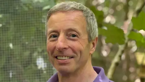 A grey haired man in his 50s wearing a purple polo shirt