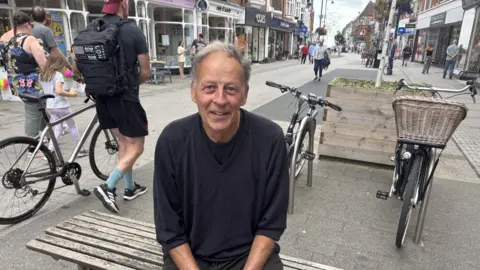 Jaimielee Rendall/BBC A man is sitting on benches in the town centre of Felixstowe. there are people walking past with bikes. He is wearing a black top and trousers.