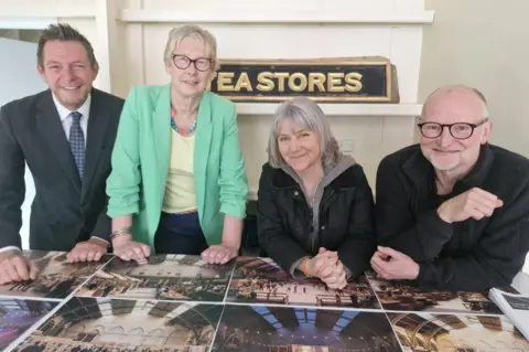 Claybody Theatre Four people are posing behind a table covered with photographs. Behind them, a vintage sign reads "TEA STORES". 