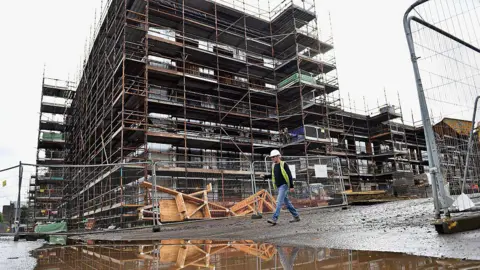 Getty Images New housing under construction in Glasgow, with a construction worker wearing jeans, a high vis jacket and a hard hat walking past a building site. Several floors of the building are under construction.