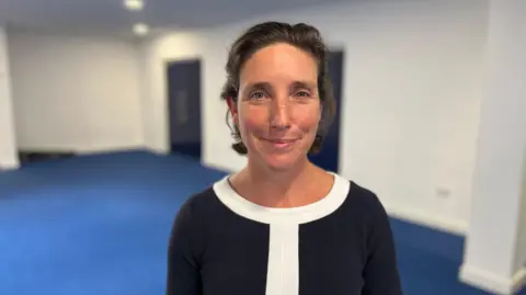 Deputy Lindsay De Sausmarez, a woman with dark hair, which is tied back from her face. She is staring at the camera. She is wearing a dark top with a round-neck, which is white. She is standing in an empty room, with a royal blue carpet and white walls. Two doors are visible behind her.