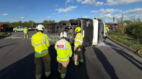 A lorry is on its side and glass is on the road. Fire crews are gathered around. A car park is in the background with police and fire vehicles.