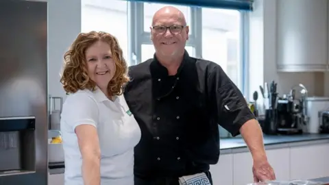 Rachael, in a white branded t-shirt, stands next to her husband Elliott who is wearing a black chef's tunic. They stand in the kitchen at their home, with a plastic box of prepared dough balls on the worktop in front of them. There is also a wooden chopping board upon which is a red pepper, brown onion and a handful of tomatoes.