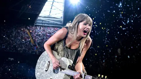 Getty Images Taylor Swift performing on stage. She is wearing a black and gold sparkly dress and is holding her silver guitar. She is looking out to the audience with dramatic facial expressions and her mouth open. Behind Swift are people sitting in the stands. The roof is closed.