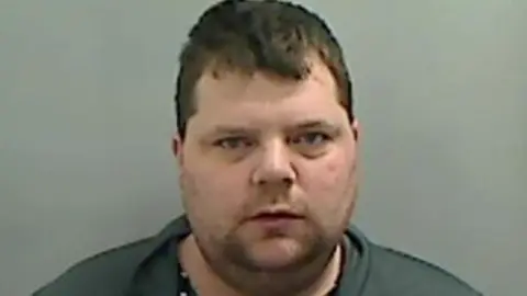 Mugshot of Alex Ibbetson. He has a large round face with faint stubble and short thick dark hair.