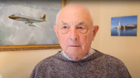 Roger Dadd - an elder gentleman wearing a speckled grey and navy jumper, with paintings of an aeroplane in the sky in a gold frame to his left and a small frameless photo of a pier on his right behind him.