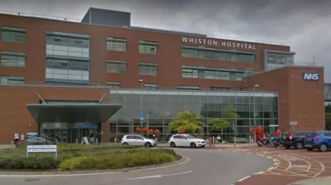 Google Exterior view of Whiston Hospital showing main entrance with a roundabout in front and a number of cars parked