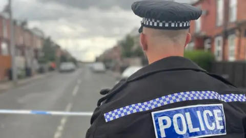 A police officer with his back to the camera wears a cap with a black and white square pattern on it and a black jacket with blue and white square edging, with the word "police" on it. He stands in front of police tape on a street