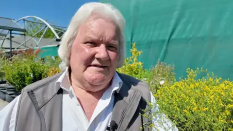 Stephen Goodridge is looking slightly off camera while giving a TV interview. He has mid length white hair and wears a white collared shirt with a grey waterproof unzipped gilet over the top. He stands in front of a row of evergreen shrubs and a green sheet in his garden centre.