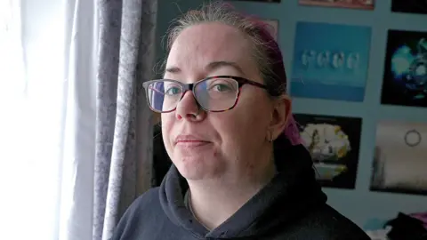 BBC Emma wears glasses and a black hoodie.