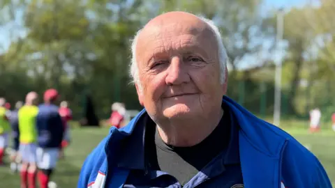 Steve Pope stood in a blue t-shirt and tracksuit top on the Astro turfed football pitch. He is smiling at the camera.
