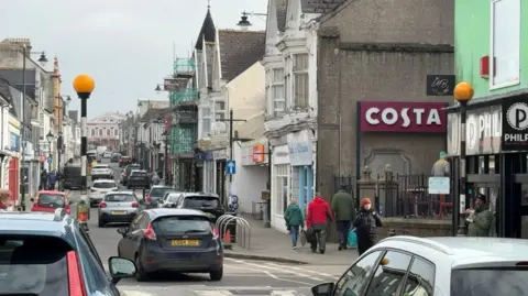 A picture taken from the end looking up at Camborne high street. There is a number of shops either side with people walking on pavements and cars driving up the street