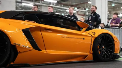 Joshua Scoot An orange supercar sits in front of a spectators who look at the vehicle. The lean over a silver barrier.