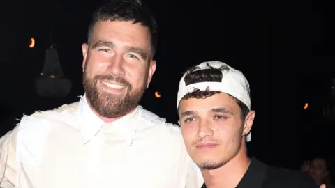 Getty Images Travis Kelce and Lando Norris at a party in Miami on 5 May. Travis is a white man in his 30s with blue eyes, short brown hair and a short brown beard. He smiles at the camera, wearing a white shirt. Lando is a 24-year-old white man with green eyes and a short moustache and goatee beard. He wears a white cap twisted back over his curly brown hair. He stands about a foot shorter than Travis, who has his arm around him. 