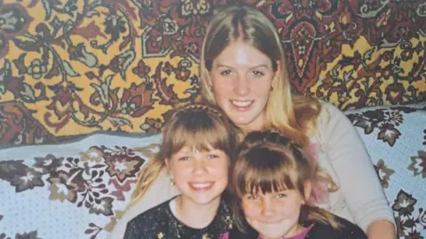 Supplied A young woman and two young girls hugging and smiling at the camera. The woman at the back has long blonde hair. The two children in front have fringes and their hair is tied up into a ponytail. They're sitting on a sofa which has a floral patterned throw over it. Behind them is a yellow, red and white tapestry. 