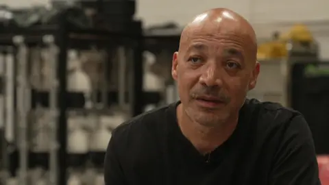 A bald black man in a black T-shirt, with brown eyes and a moustache sits in front of a bank of CCTV cameras hanging up behind him