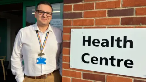 David Lewis is standing in front of the Beccles Medical Centre. He is wearing a white shirt and light brown trousers. He has a rainbow lanyard and name badge. He is smiling and wearing glasses. To the right of where Mr Lewis is standing is a sign which says Health Centre in black on a white background. 