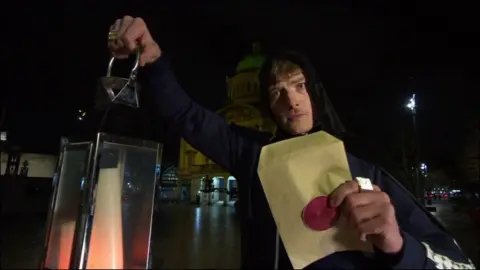 Jack Marriner-Brown poses in Hull city centre wearing a black hooded cloak. The Traitors contestant is holding a lantern containing a candle and an envelope sealed with wax. Hull City Hall is visible in the background.