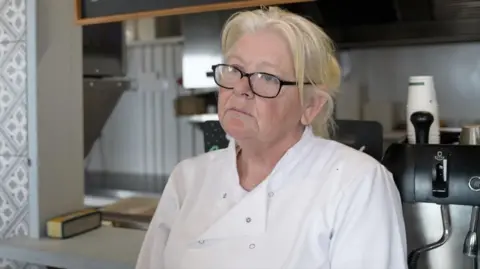 Blonde woman with glasses wearing white overalls in a kitchen
