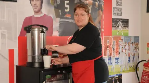 A woman with mousey brown hair and a fringe with her hair in a ponytail. She is wearing glasses and smiling at the camera while making a hot drink at a machine. She is wearing a red apron and black top and football photos are on the walls.