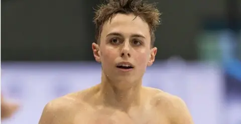Getty Images Filip Nowacki is bare-chested and has wet hair and shoulders. He is standing in front of a blurred pool