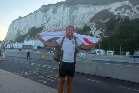 James Lewis A smiling James Lewis wears a white England top and dark shorts. He is standing in front of the white cliffs of Dover and holding a flag aloft. 