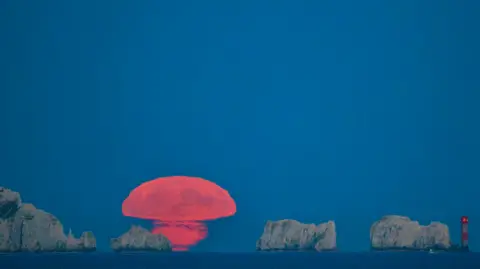Gary Jacobs The moon above a rock and cliff formation off the Isle of Wight, a red and white lighthouse to the right. The moon is melding into the horizon like an orange mushroom.