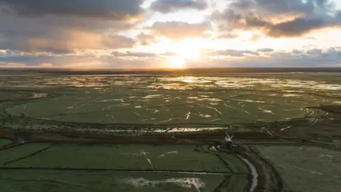 Broads Authority Aerial image of Halvergate marshes during a winter sunrise, showing Mutton's Mill (windmill) in the foreground and a waterlogged flat green landscape.