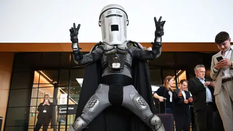 Getty Images A man with a bin on his head poses triumphantly outside a giant glass window. The bin is silver with a slit cut out for his eyes and a square hold for his mouth. He also wears black gloves, a black breastplate, knee and elbow guards, and a long silver cape. 